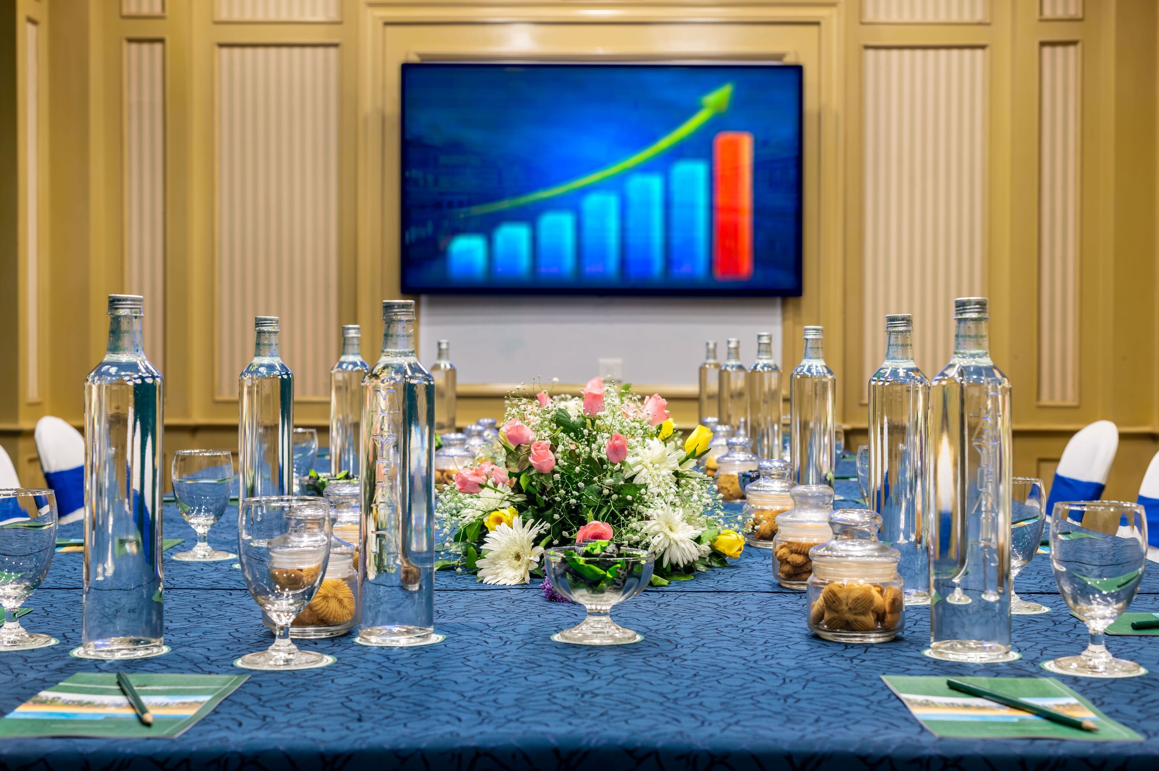 A conference table set with clear glass bottles, a floral centerpiece, and a television displaying a bar graph.