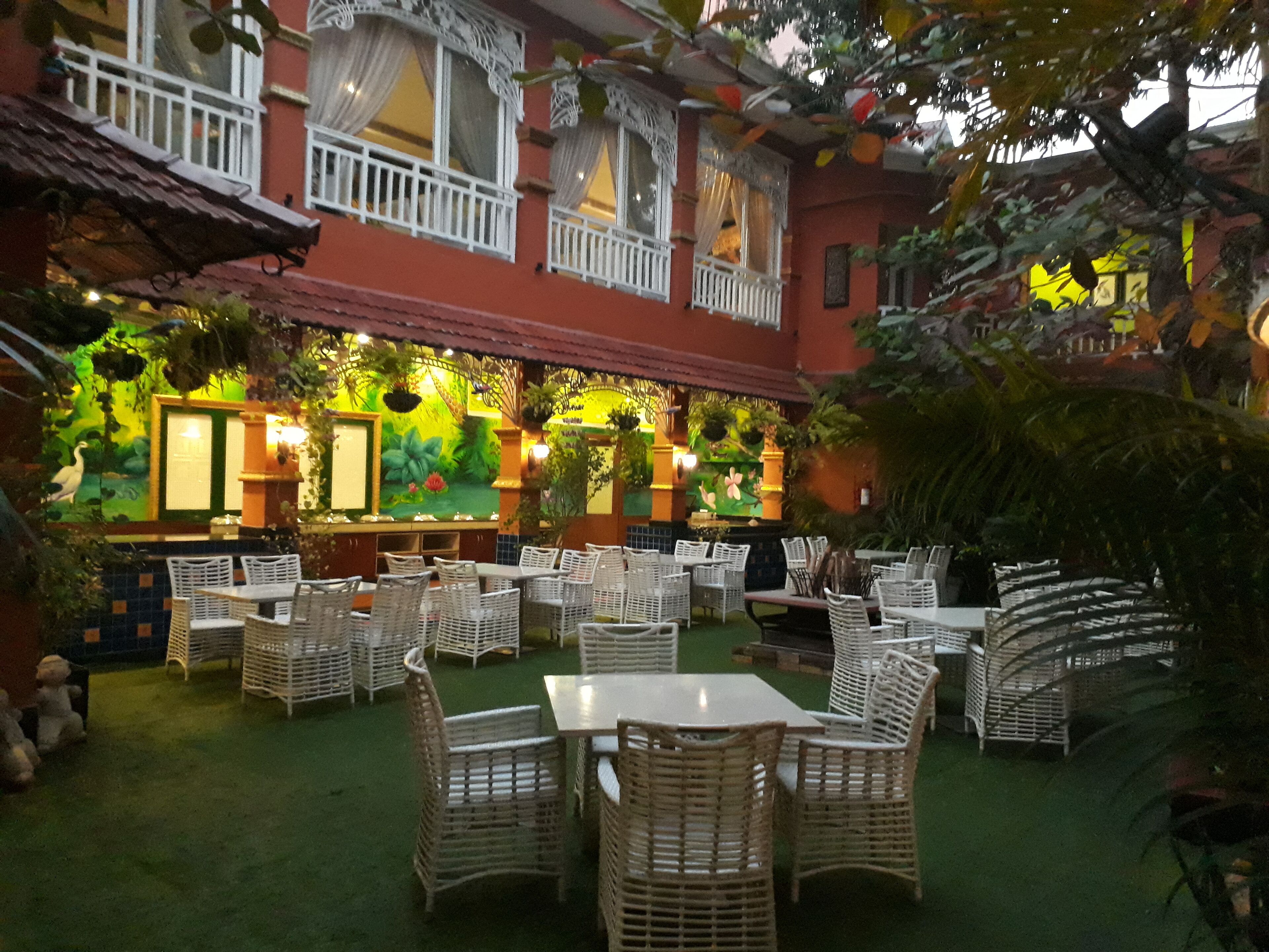 An outdoor dining area with wicker tables and chairs, a mural on the wall, and a balcony with white railings.