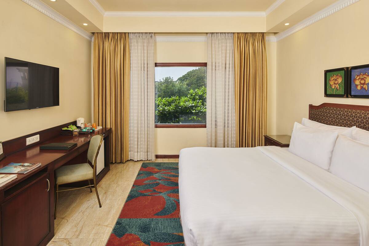 Habitación de hotel con cama, escritorio, silla, televisor, ventana con cortinas y una obra de arte en la pared.