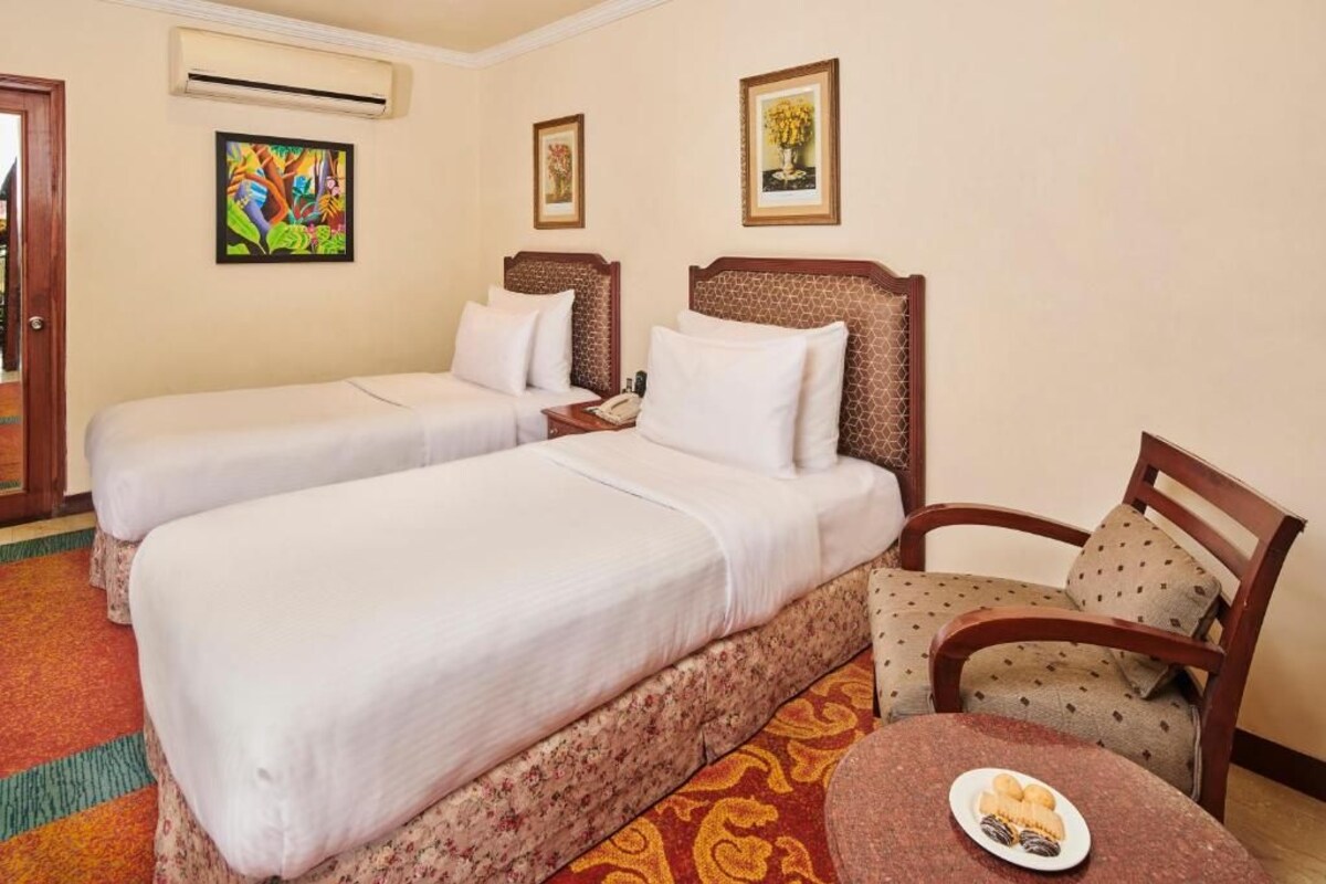Une chambre d’hôtel avec deux lits, une chaise, une petite table sur laquelle se trouve une assiette de nourriture et un tableau accroché au mur.