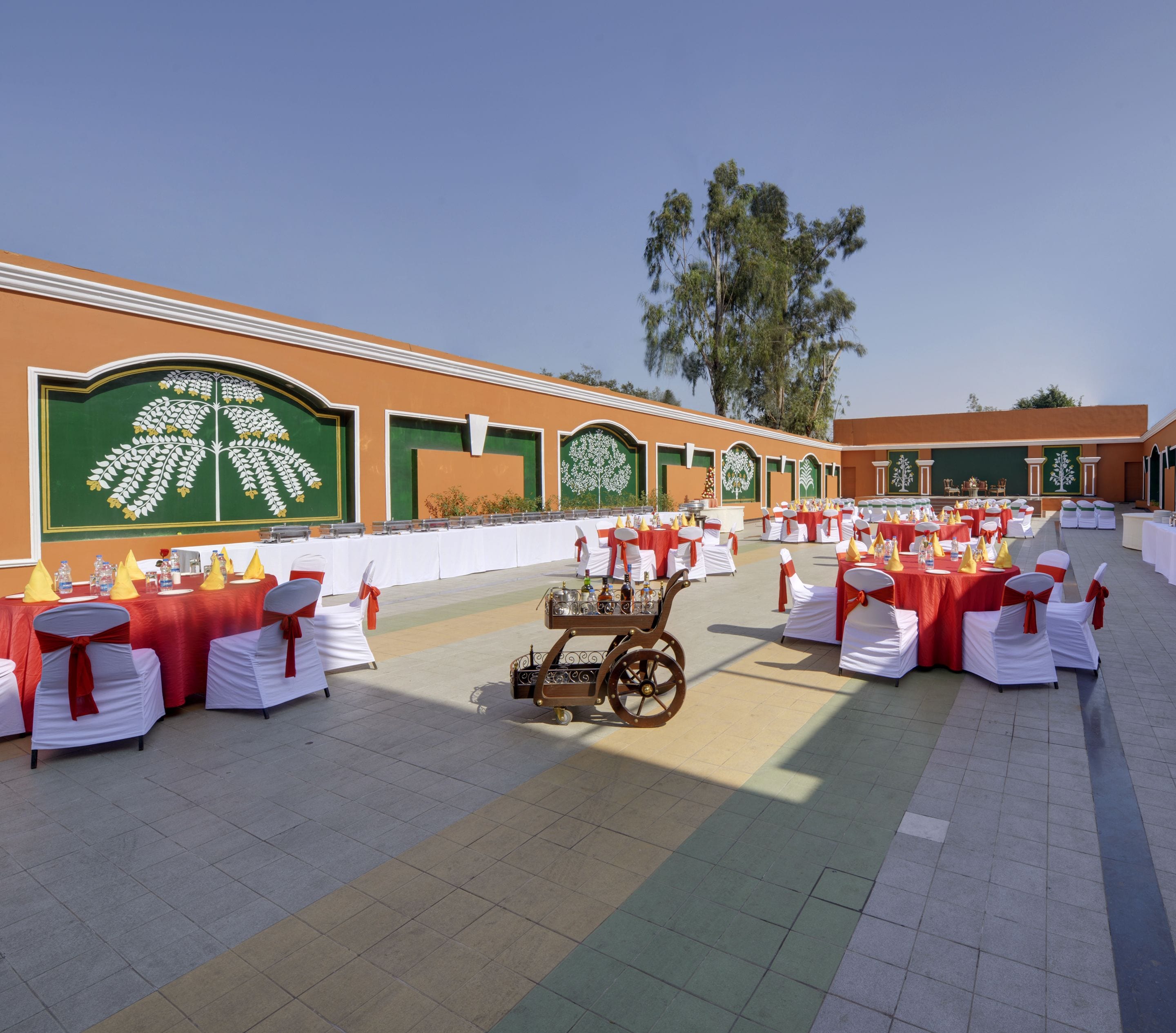 Un salón de banquetes al aire libre con filas de mesas dispuestas para comer, un carro con cristalería y un fondo decorado con diseños florales en tonos verdes y blancos.
