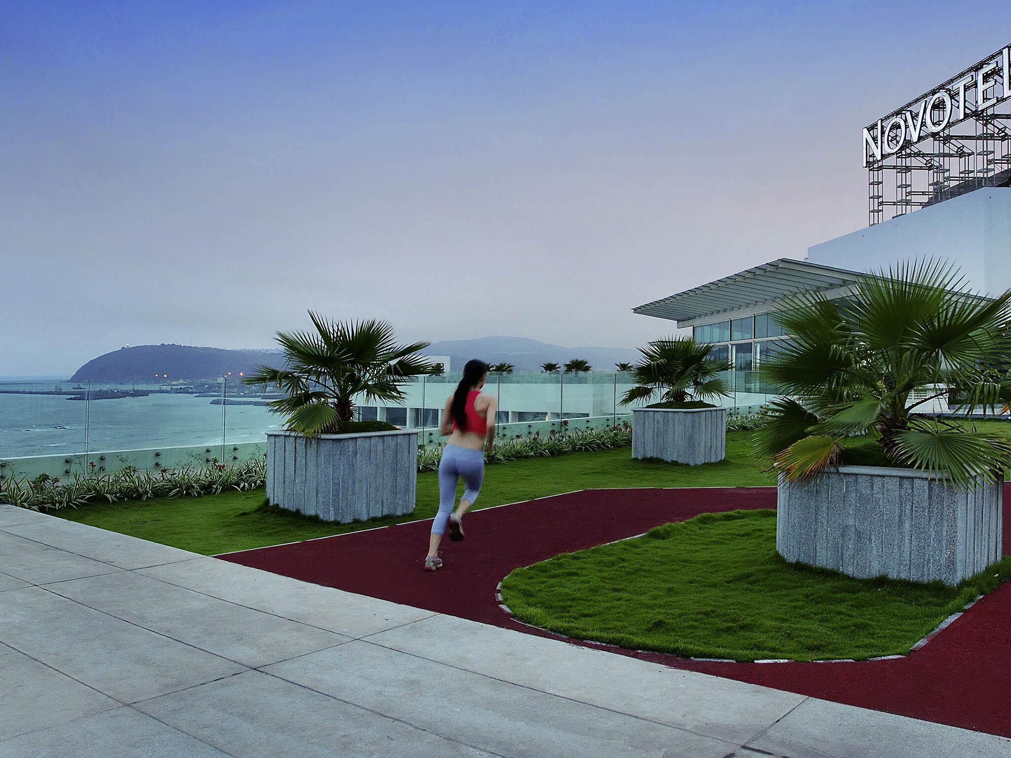 A modern hotel with a view of the sea, featuring a red running track, palm trees, and a sign reading "NOVOTEL".