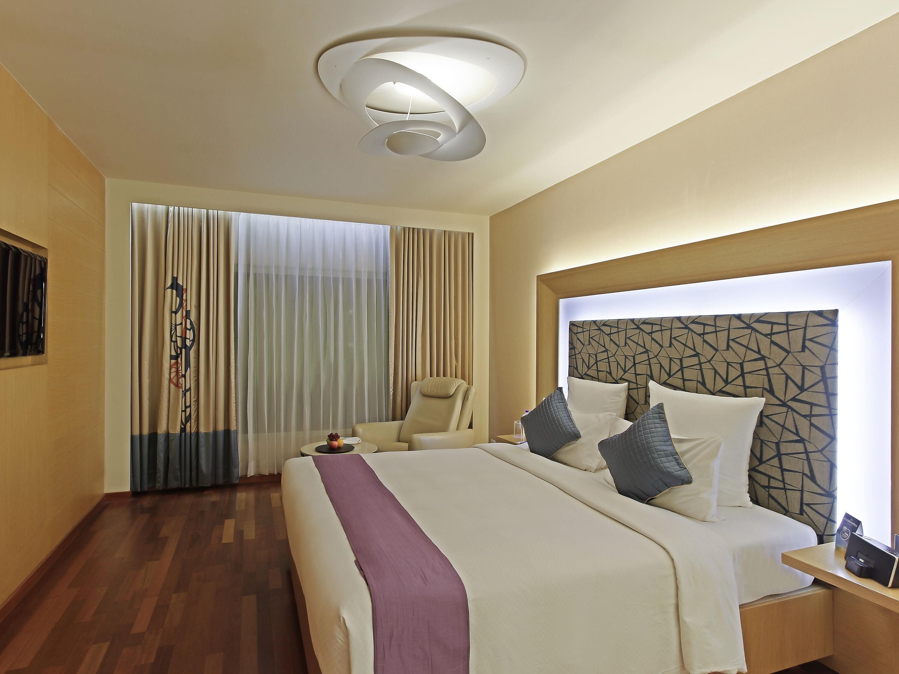 A hotel room with a large bed, a chair, a bedside table, and a modern ceiling light fixture.