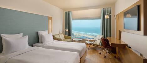 A hotel room with two beds, a desk, a chair, a small table, and a view of the beach.