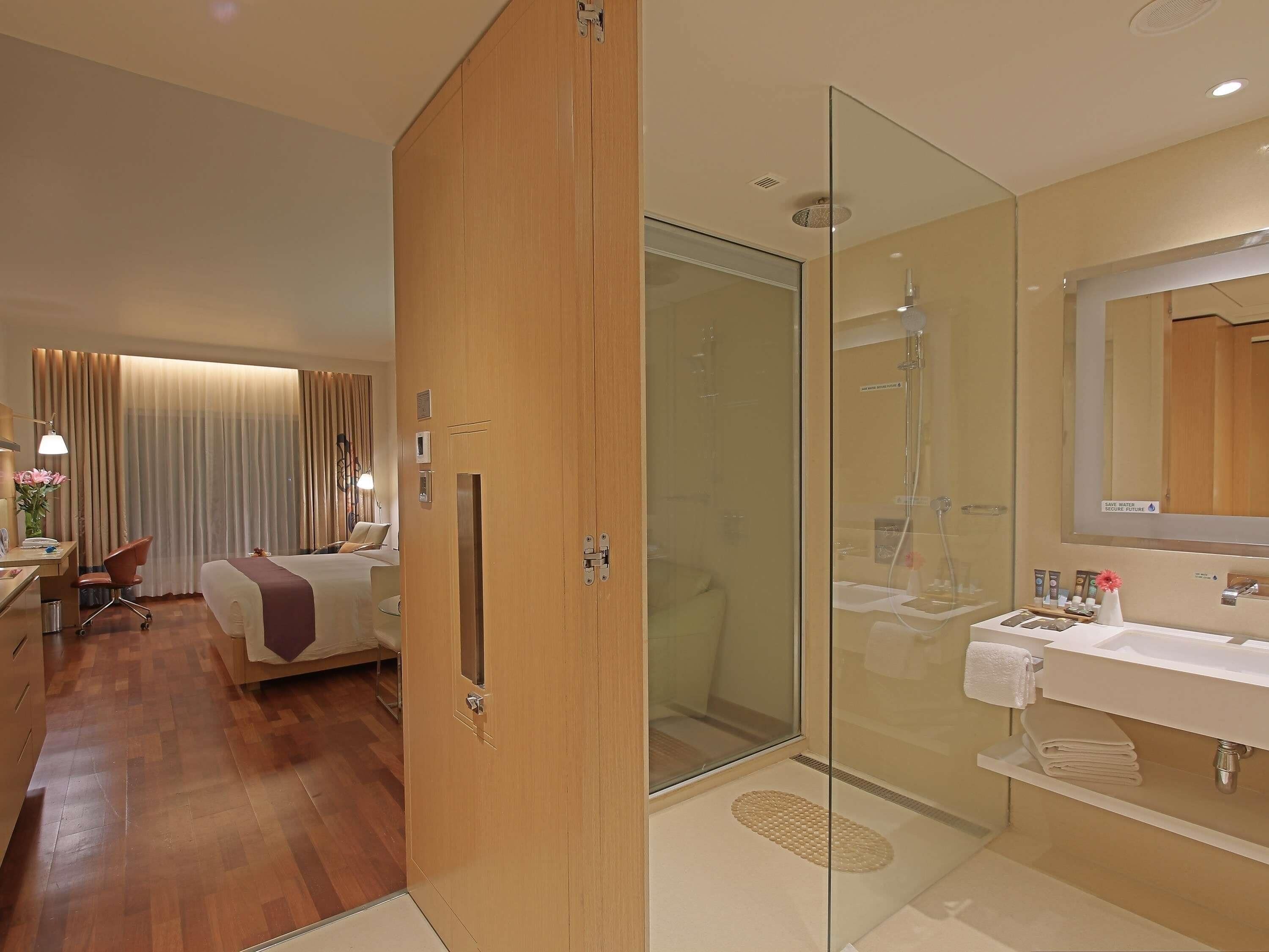 Habitación de hotel con armario de madera, cama, escritorio con silla, mesita de noche, ducha con cerramiento de vidrio, lavamanos blanco y espejo.