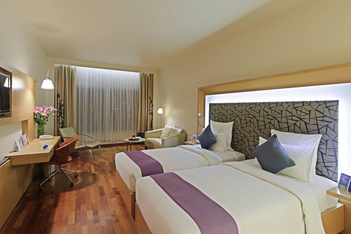 Une chambre d’hôtel avec deux lits, un bureau avec une chaise, une petite table et une grande fenêtre avec des rideaux.