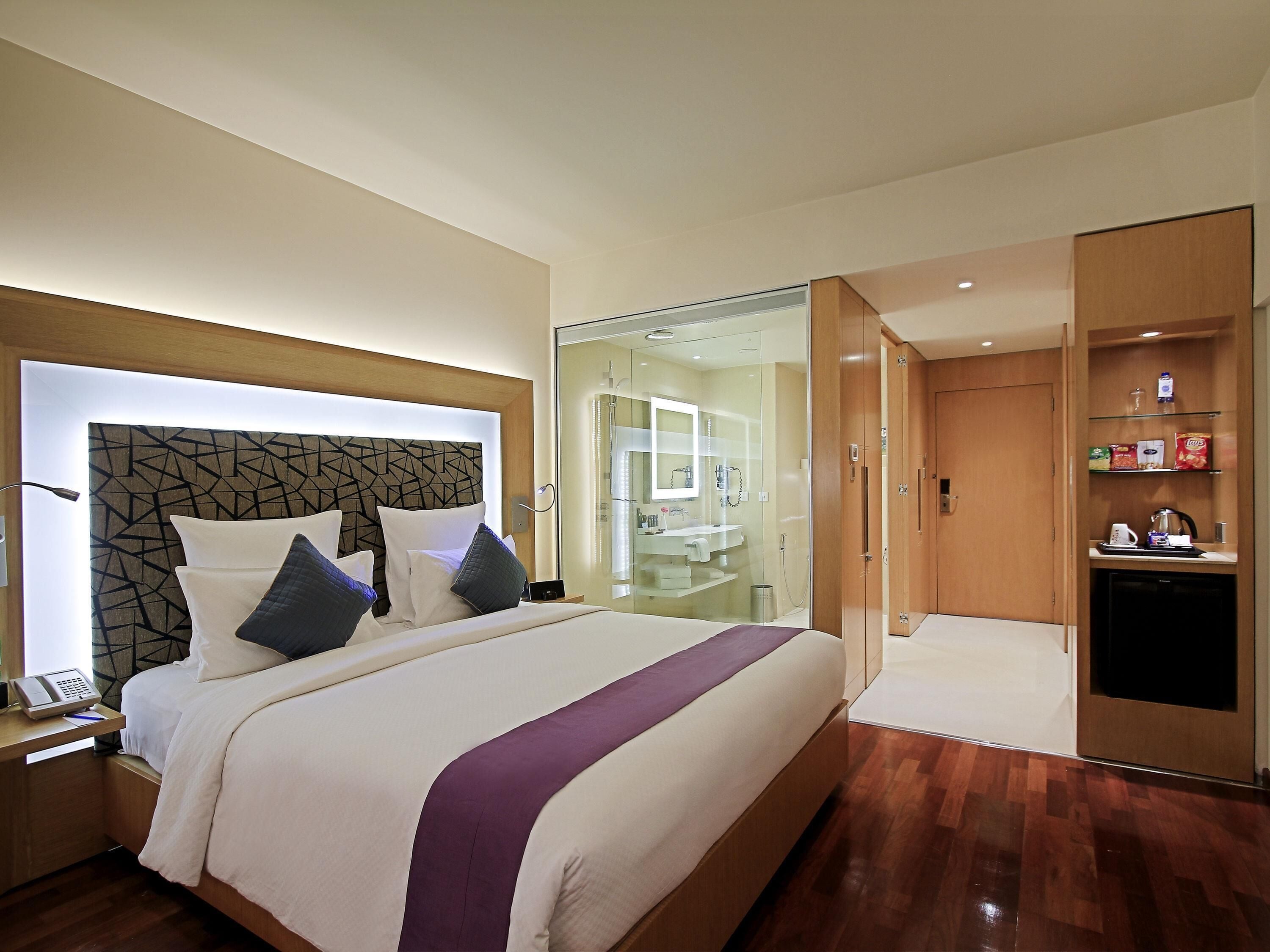 A modern hotel room with a large bed, a bedside table with a phone, a glass-enclosed shower area, and a wooden wardrobe with shelves.