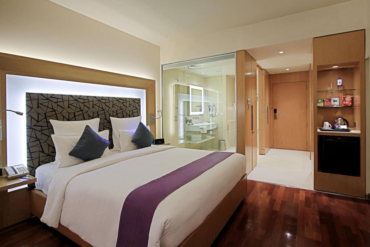 Ein modernes Hotelzimmer mit einem großen Bett, einem Nachttisch mit Telefon, einer gläsernen Duschkabine und einem Holzkoffer mit Ablagen.