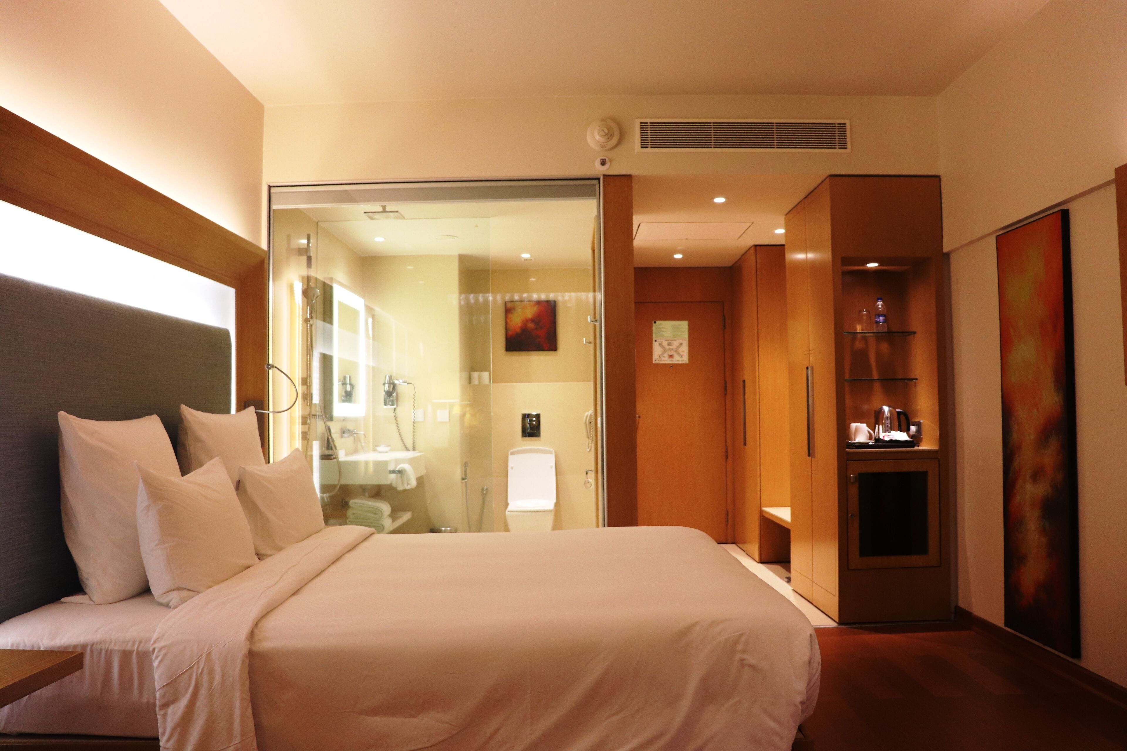 A hotel room with a large bed, a glass shower enclosure, a small cabinet, and a painting on the wall.