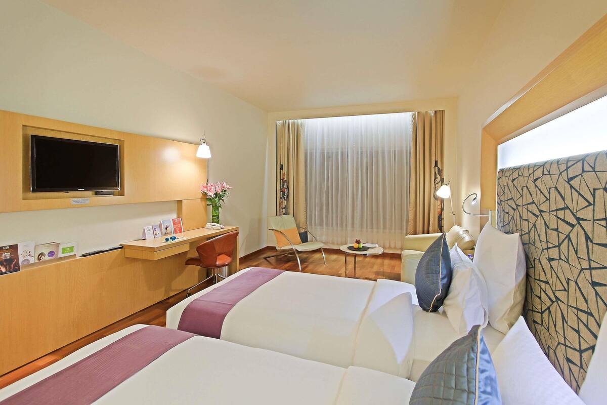 A hotel room with two beds, a desk with a chair, a TV mounted on the wall, and a large window with curtains.