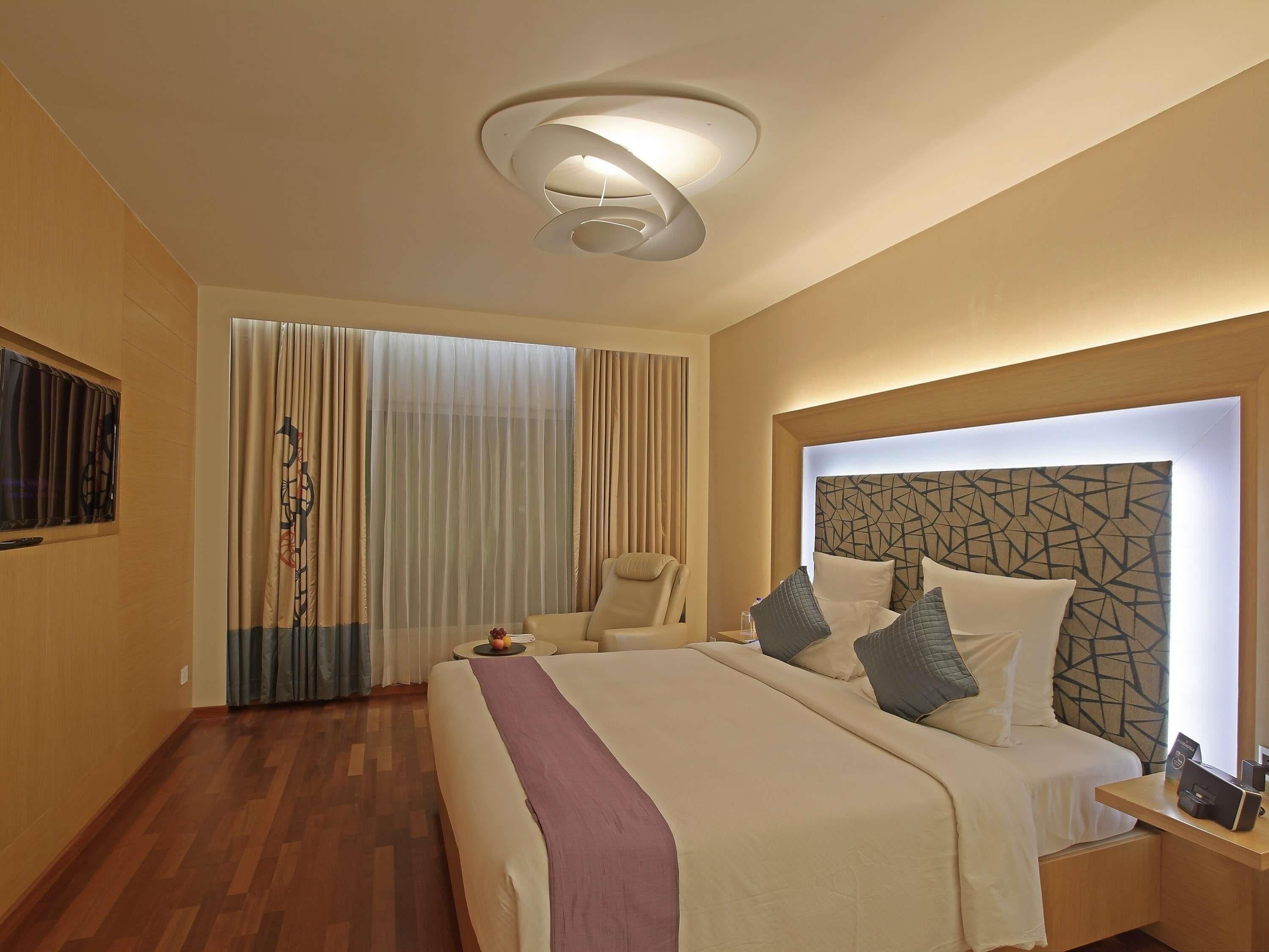 A hotel room with a bed, a chair, a desk, a wall-mounted TV, and a ceiling light fixture.