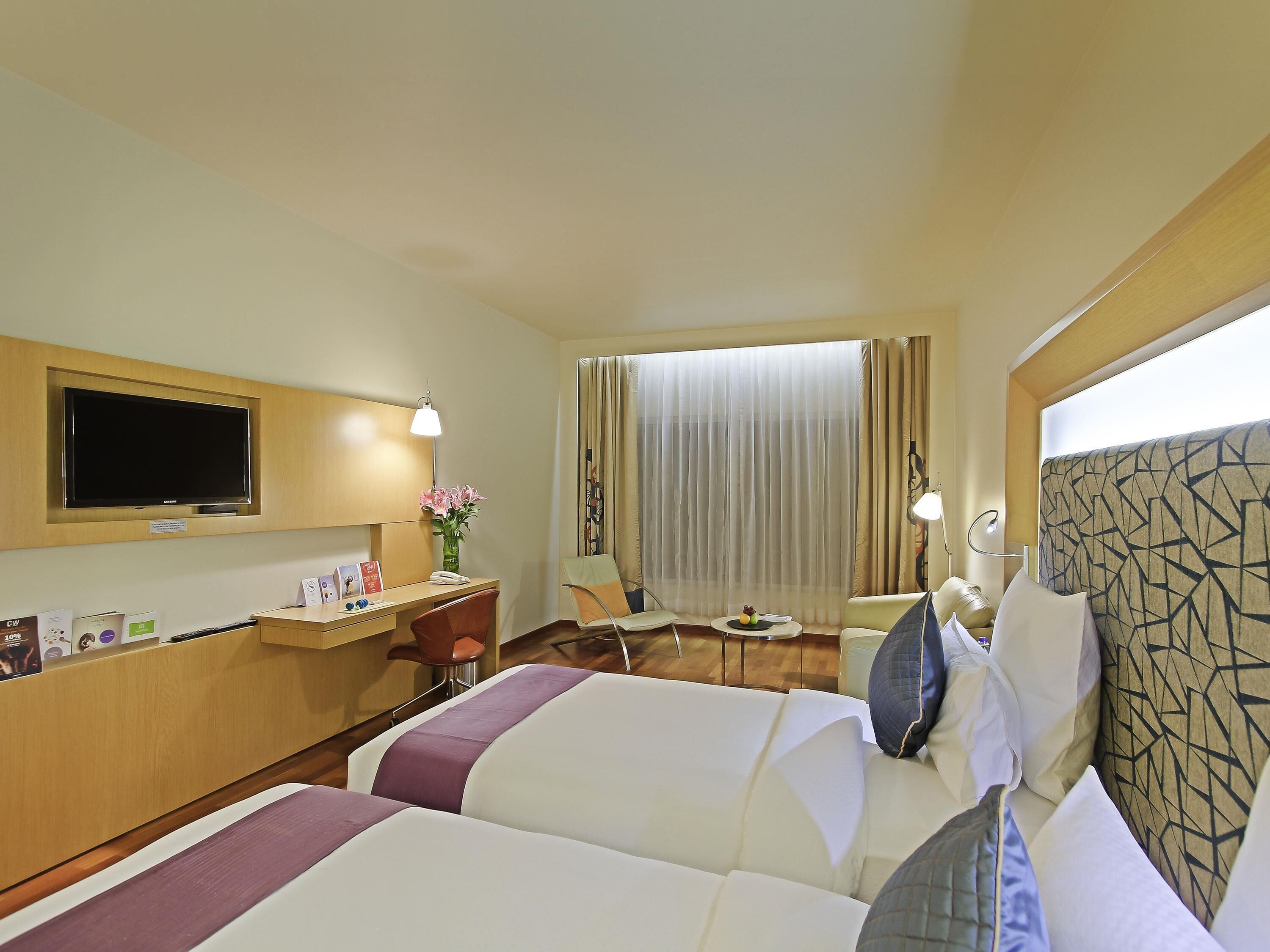 A hotel room with two beds, a desk, a TV, and a window with curtains.