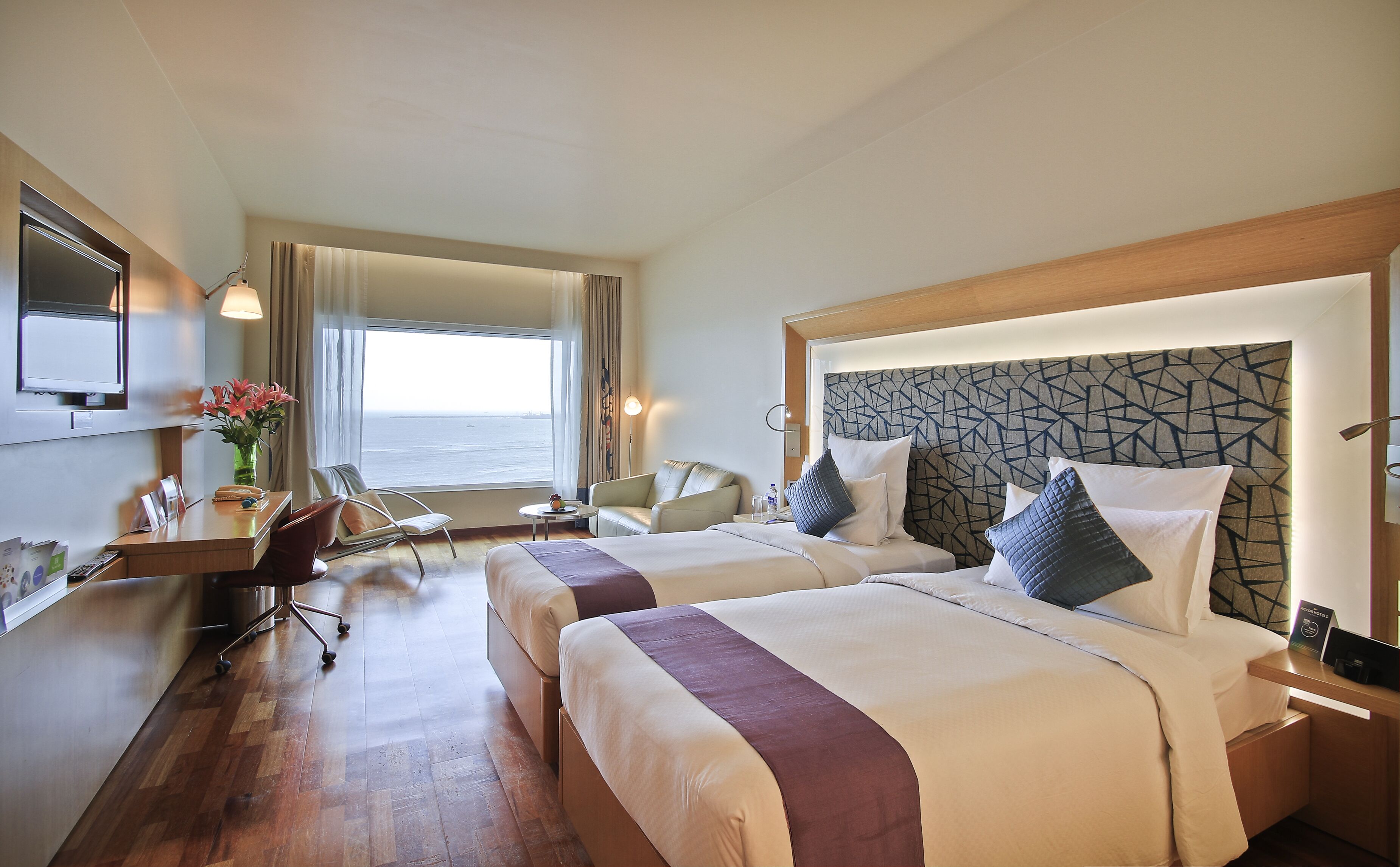 Quarto de hotel com duas camas, uma escrivaninha, uma cadeira, uma televisão e vista para o mar.