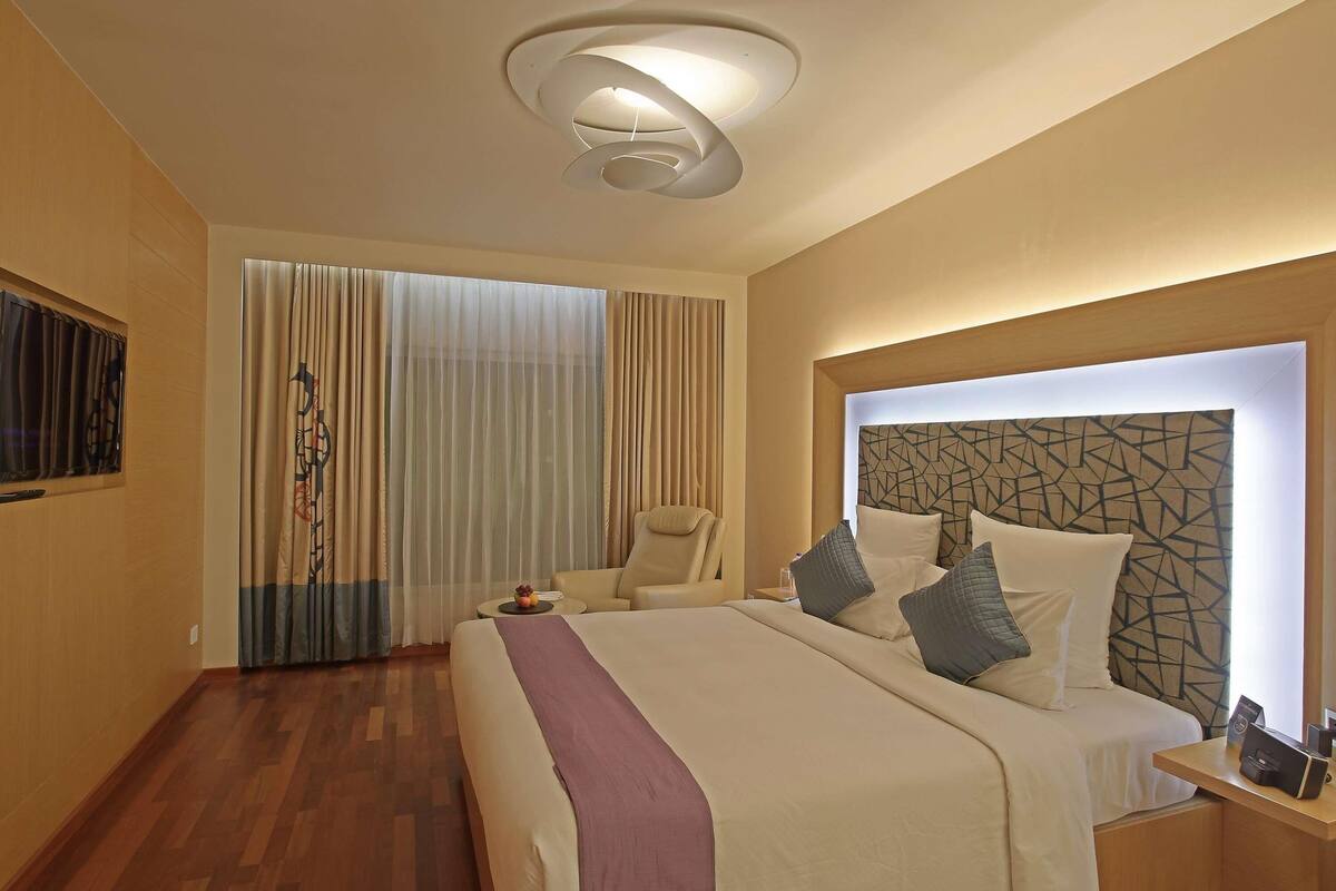 A hotel room with a bed, a chair, a desk, a wall-mounted TV, and a ceiling light fixture.