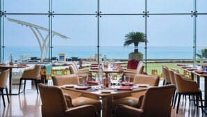 A restaurant with tables set for dining, large windows offering a view of the sea, and outdoor seating with chairs and tables. - Novotel Visakhapatnam Varun Beach Hotel (Visakhapatnam)