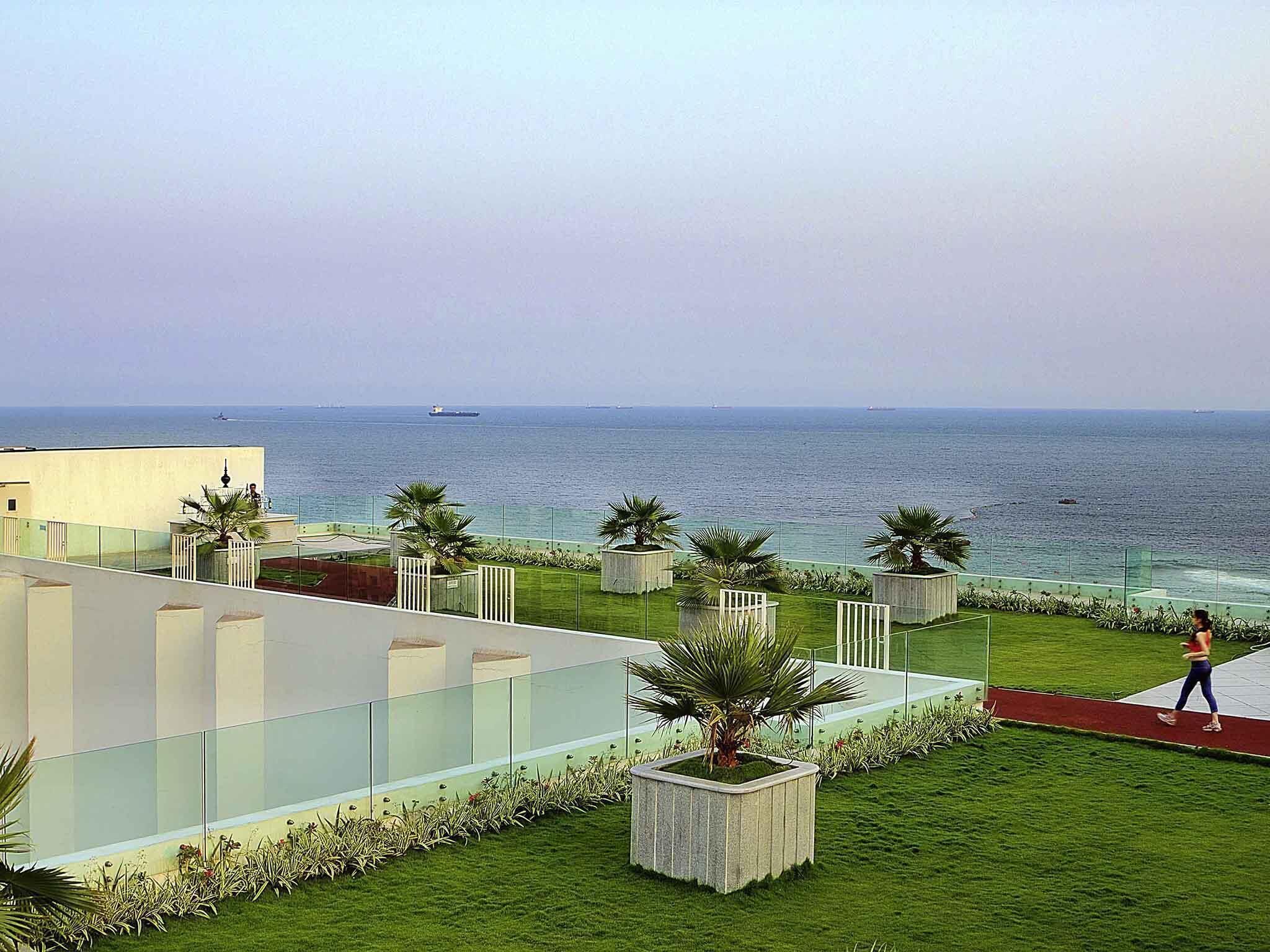 Un edificio moderno con fachada de vidrio que ofrece vistas al mar. Se observan palmeras y un sendero rojo.