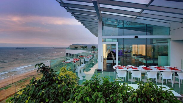 A modern building with a glass facade overlooking the ocean, featuring outdoor seating and a bar area. - Novotel Visakhapatnam Varun Beach Hotel (Visakhapatnam)