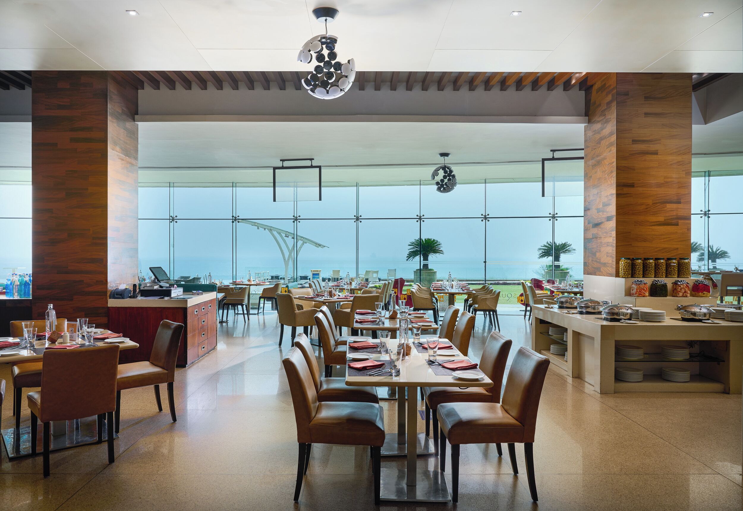 Un comedor amplio con varias mesas y sillas, ventanas grandes que ofrecen vistas al océano y un mostrador de buffet con diversos alimentos.