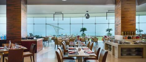 Un comedor amplio con varias mesas y sillas, ventanas grandes que ofrecen vistas al océano y un mostrador de buffet con diversos alimentos.