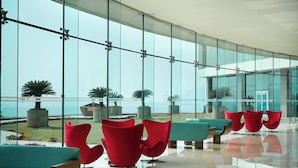 A modern lobby with large glass windows, red and teal seating, and a view of the sea. - Novotel Visakhapatnam Varun Beach Hotel (Visakhapatnam)