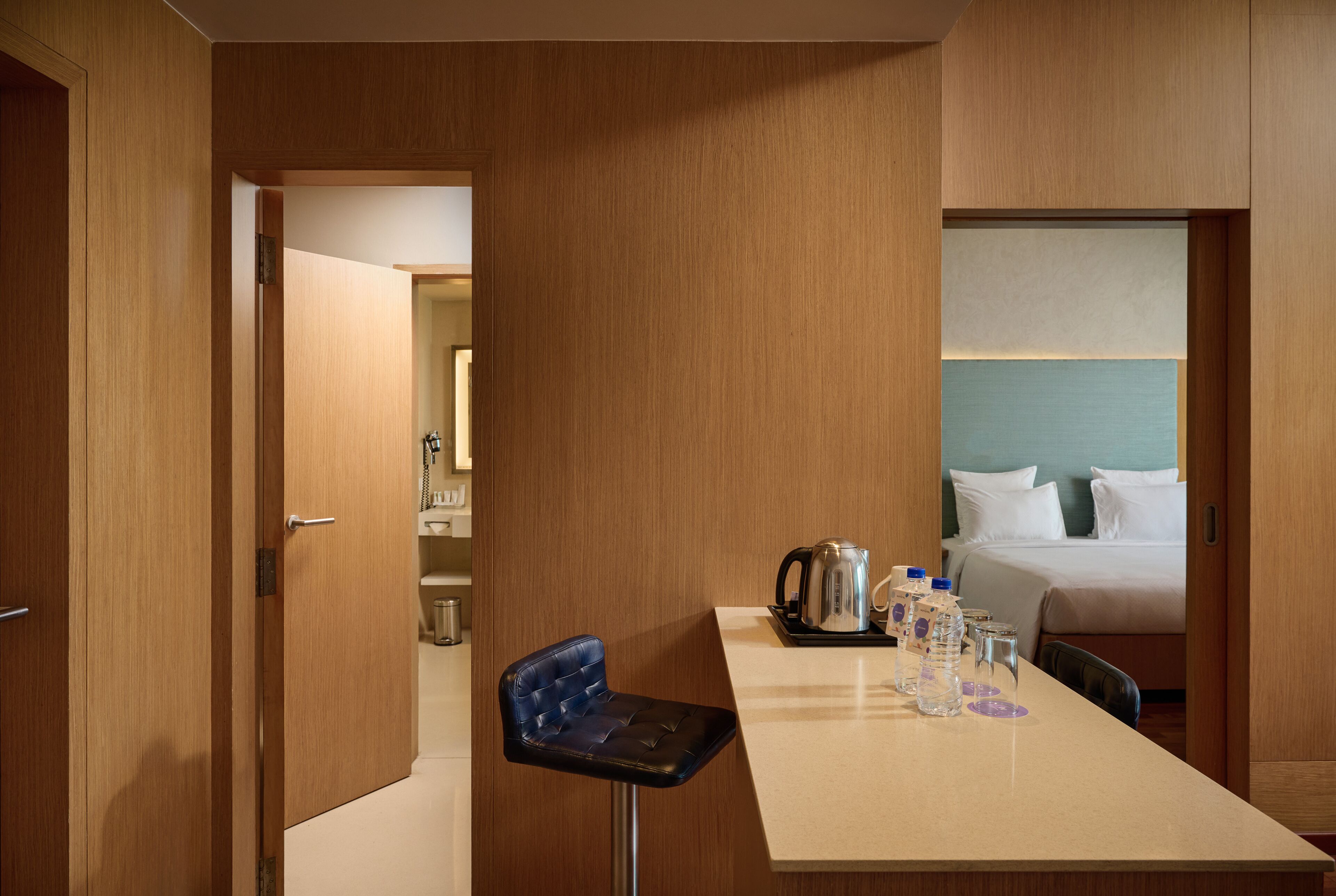 A hotel room with a wooden door, a kitchenette, a bed with white bedding, and a small bar area with a blue bar stool.