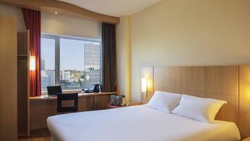 Habitación de hotel con una cama grande, un escritorio con una computadora portátil, una cafetera y vistas a un paisaje urbano a través de un ventanal amplio.