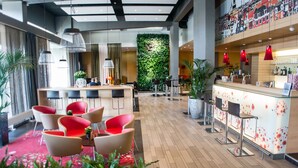 A modern hotel lobby with a reception desk, red and white chairs, a green vertical garden, and a bar area with red pendant lights. - ibis Leiden Centre (Leiden)