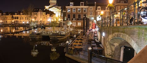 Vista nocturna de un canal con barcos amarrados, un puente y edificios con ventanas iluminadas.