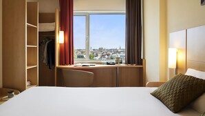 A hotel room with a bed, a desk with a chair, a wardrobe, and a large window offering a city view. - ibis Leiden Centre (Leiden)