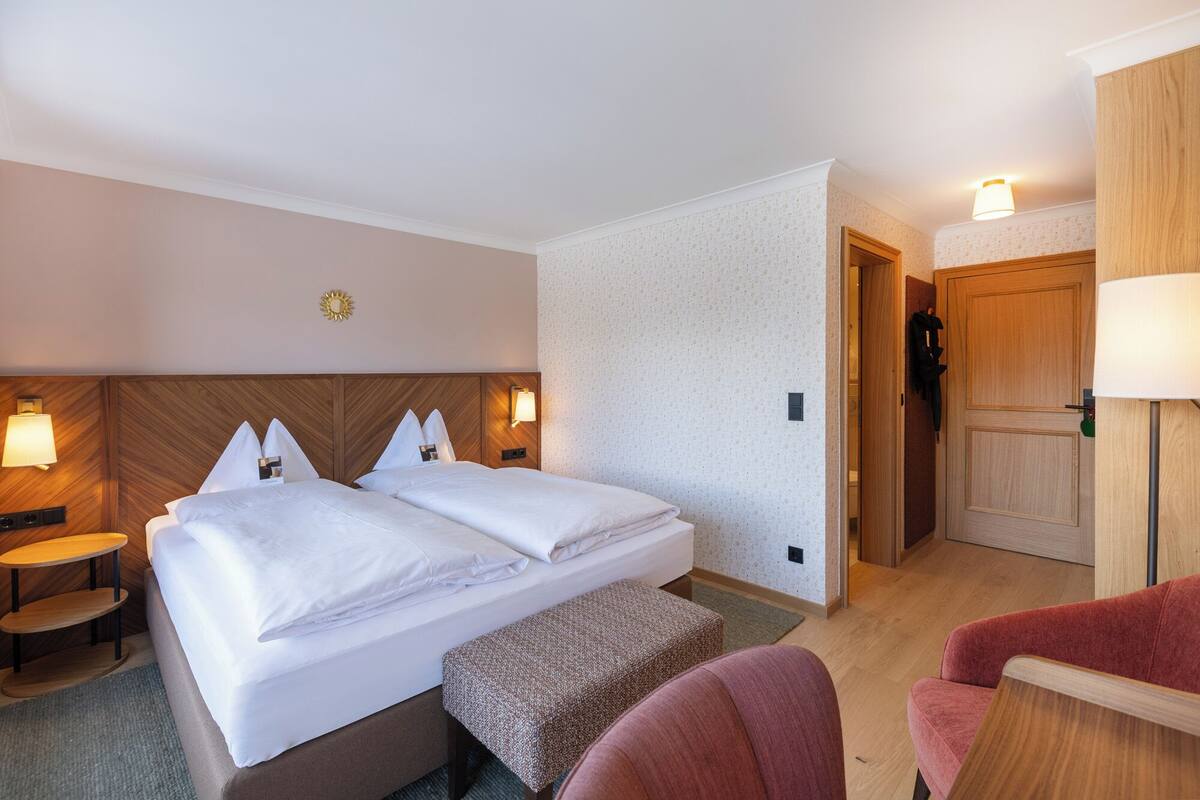 A hotel room with a double bed, bedside lamps, a small table, a bench, and a door leading to a bathroom.