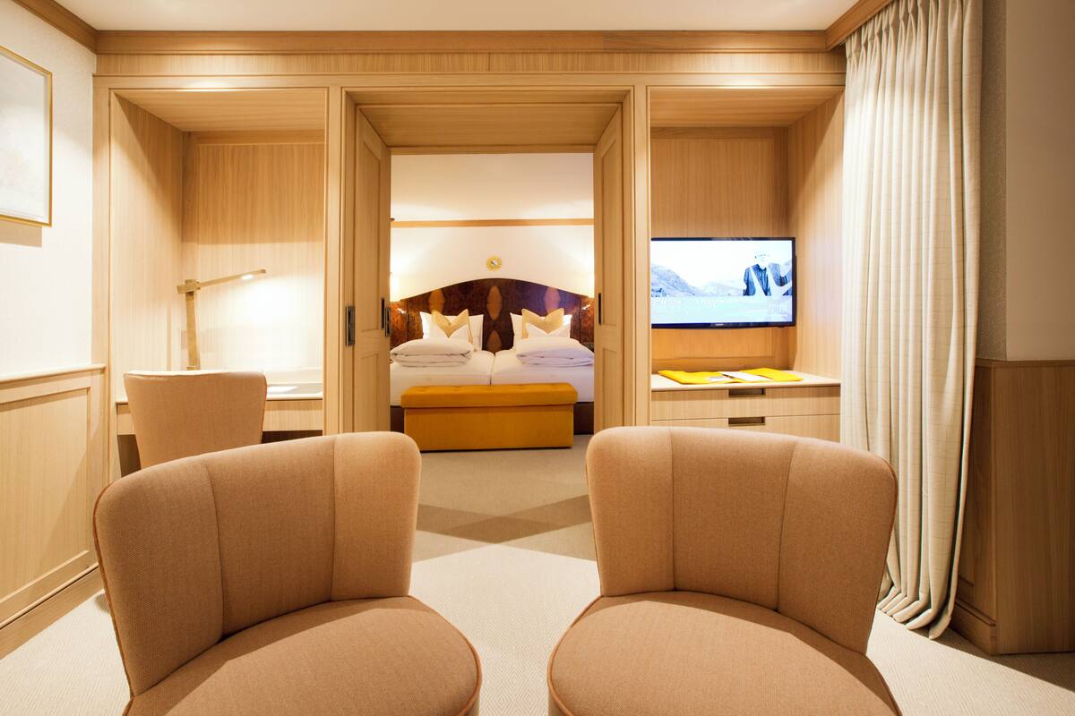A hotel room with a bed, two armchairs, a desk, a TV, and a large window.