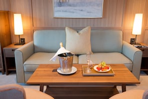 A hotel room with a light blue sofa, a wooden coffee table with a champagne bucket and fruit tray, and two bedside lamps.