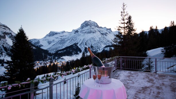 Una mesa con una botella de champán y dos copas en un balcón con vista a una montaña nevada.