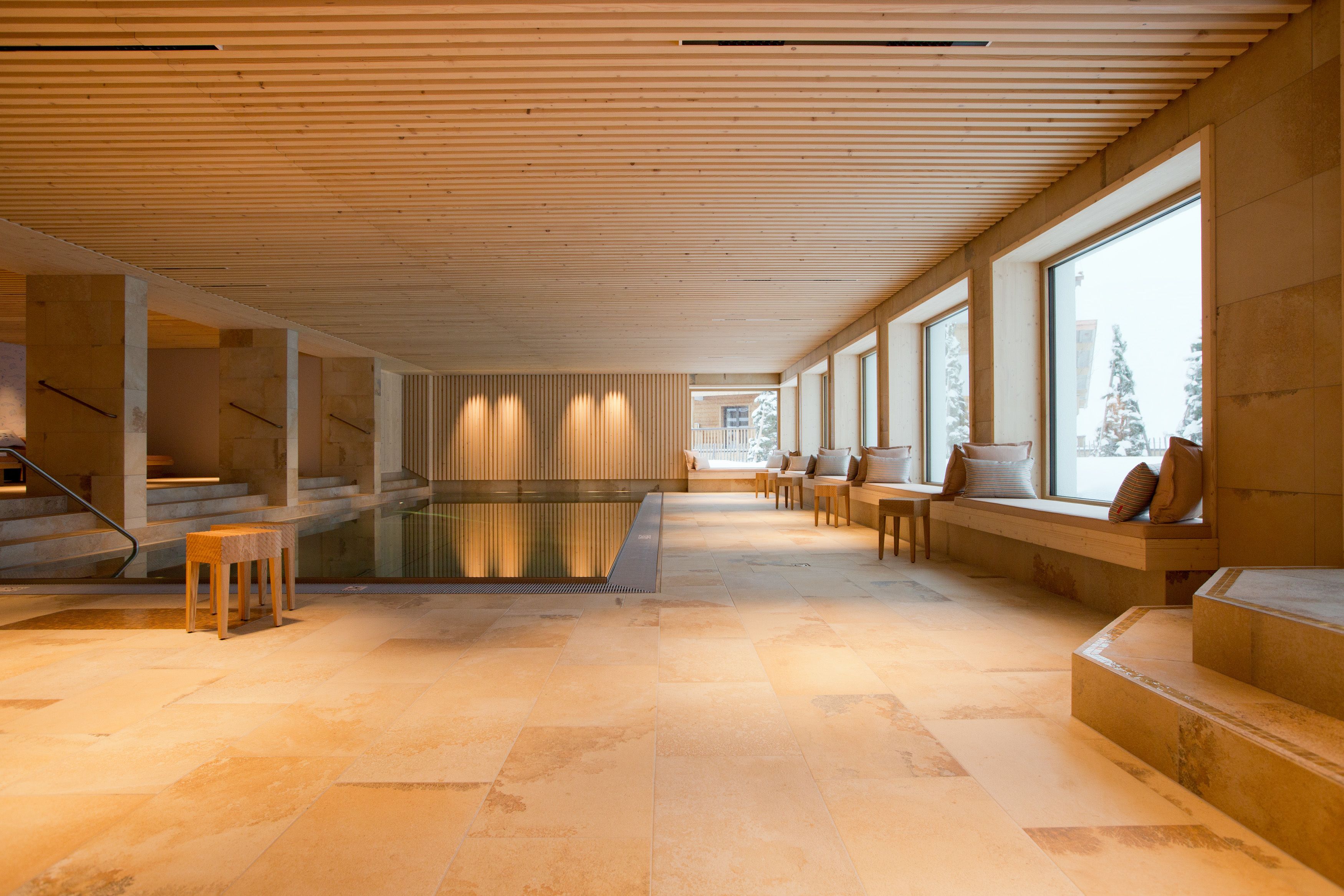 A spacious indoor area with a pool, wooden beams, large windows, and seating along the walls.