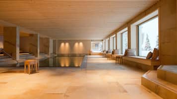 A spacious indoor area with a pool, wooden beams, large windows, and seating along the walls.