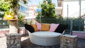 A round wicker sofa with cushions, two wooden stools, and a metal fence in an outdoor setting. - Hotel Angelini (Rimini)