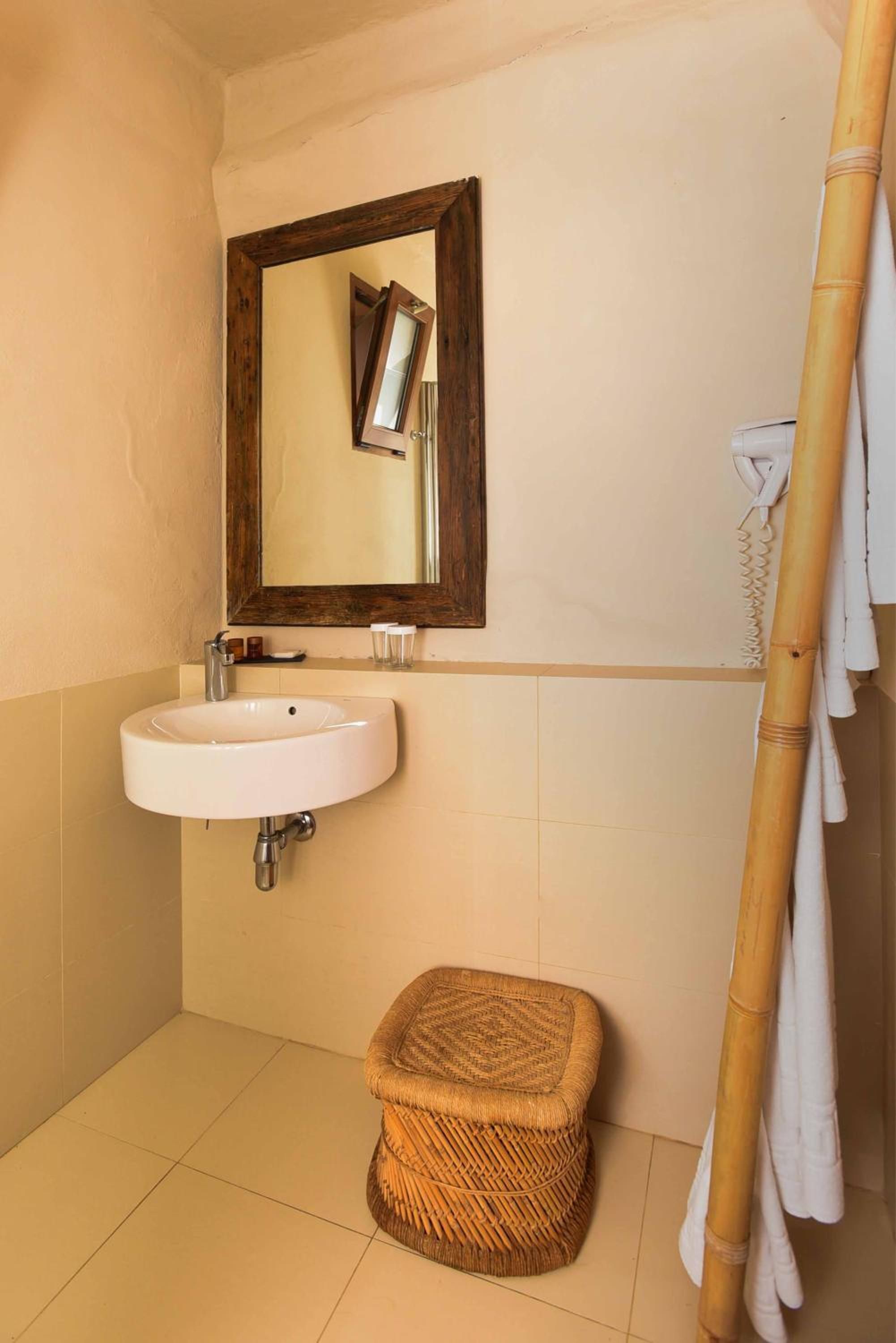 A bathroom with a white sink, a wooden-framed mirror, a wicker stool, and a bamboo towel rack.