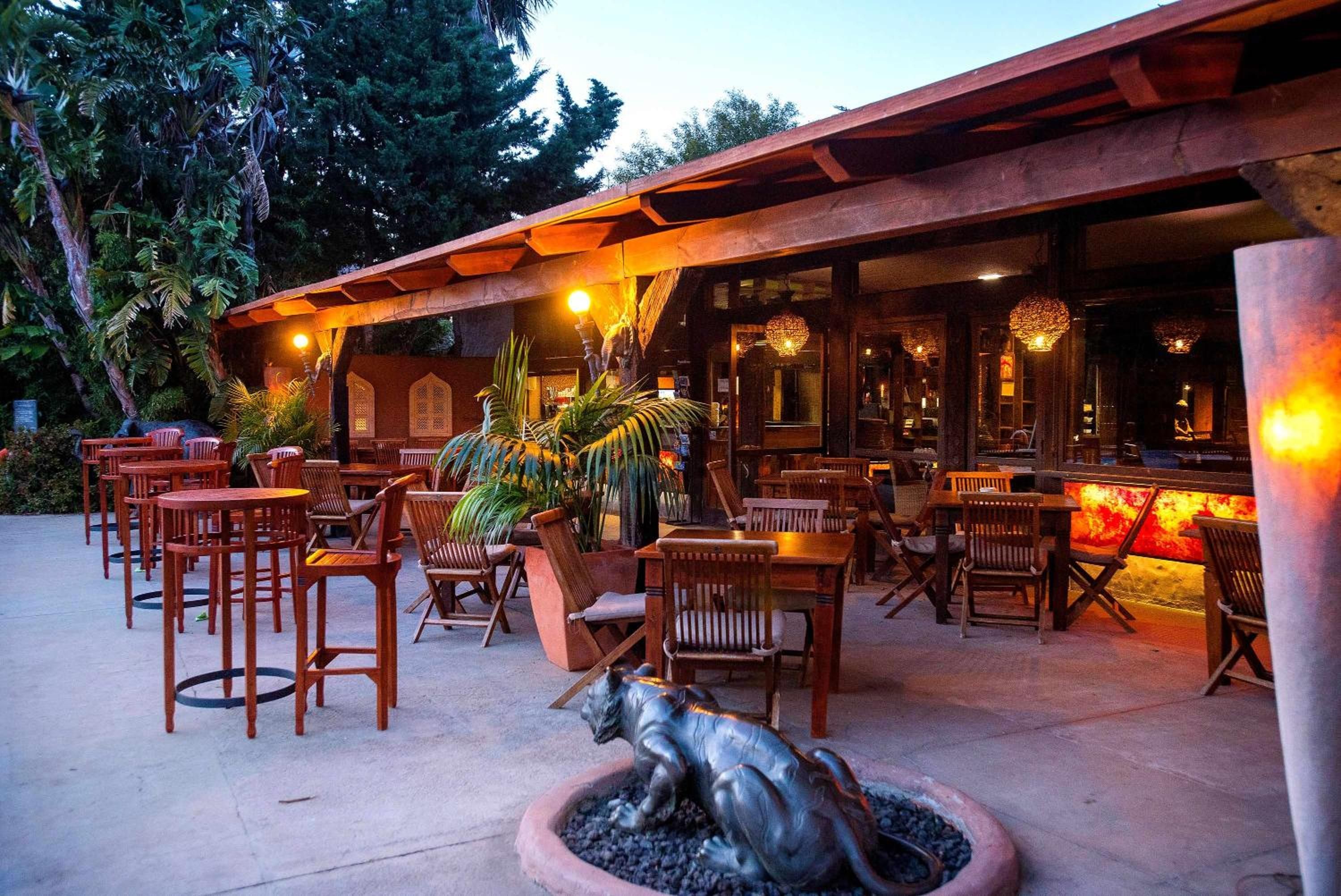 An outdoor seating area with wooden tables and chairs, surrounded by tropical plants and a decorative dog statue.