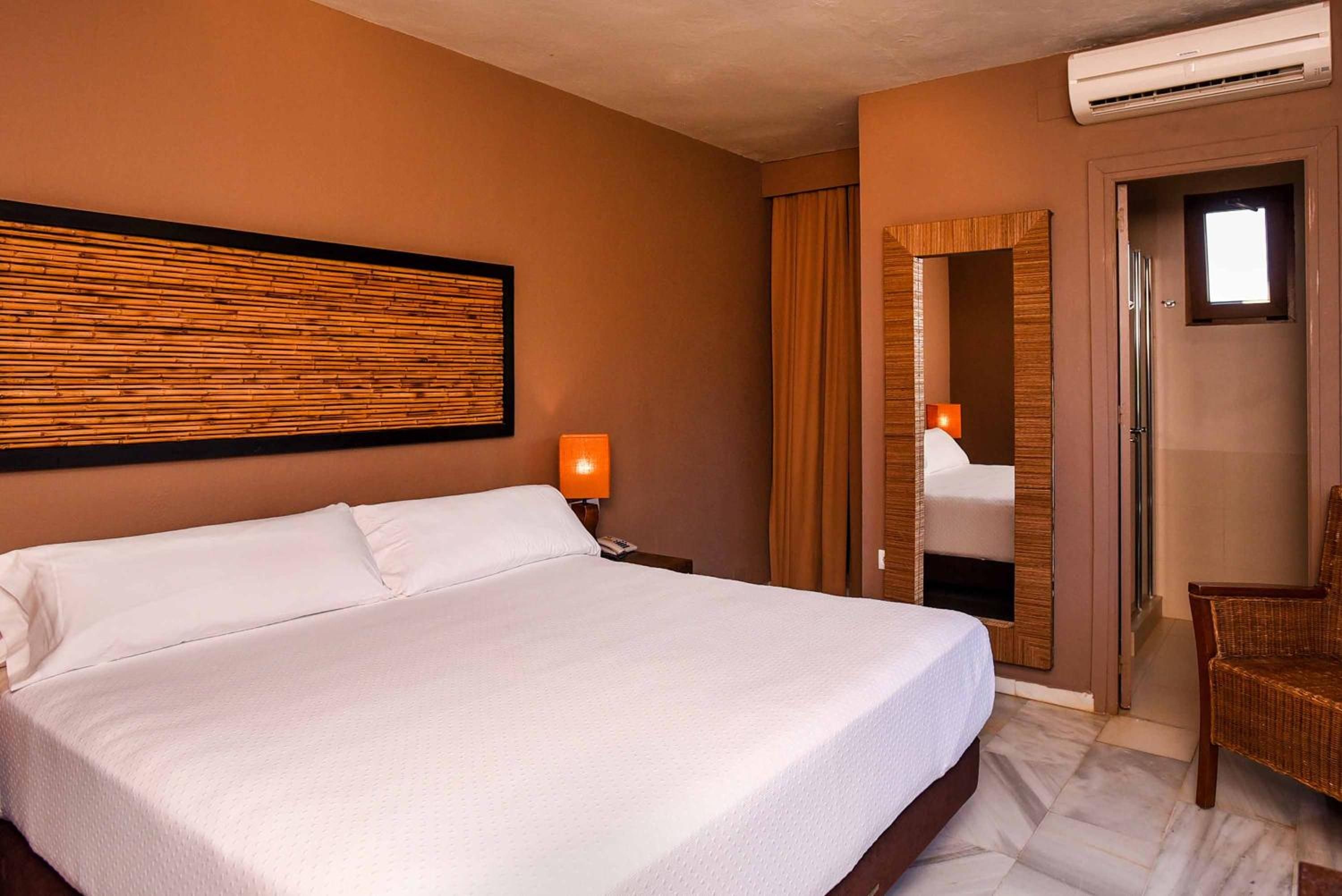 A hotel room with a large bed, a nightstand with a lamp, a mirror, and a wicker chair.