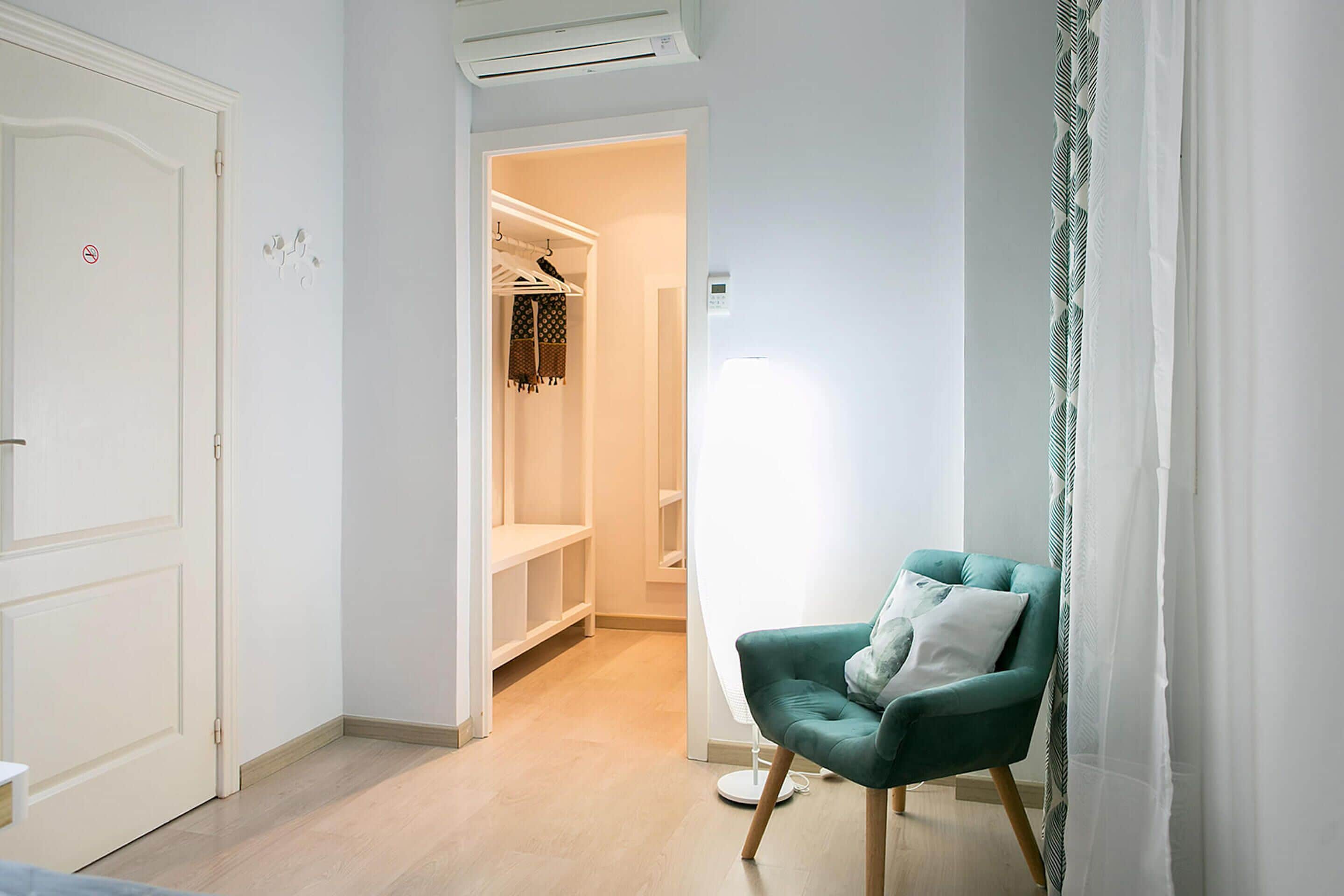 A hallway with a green armchair, a white door, and a closet with hanging clothes.