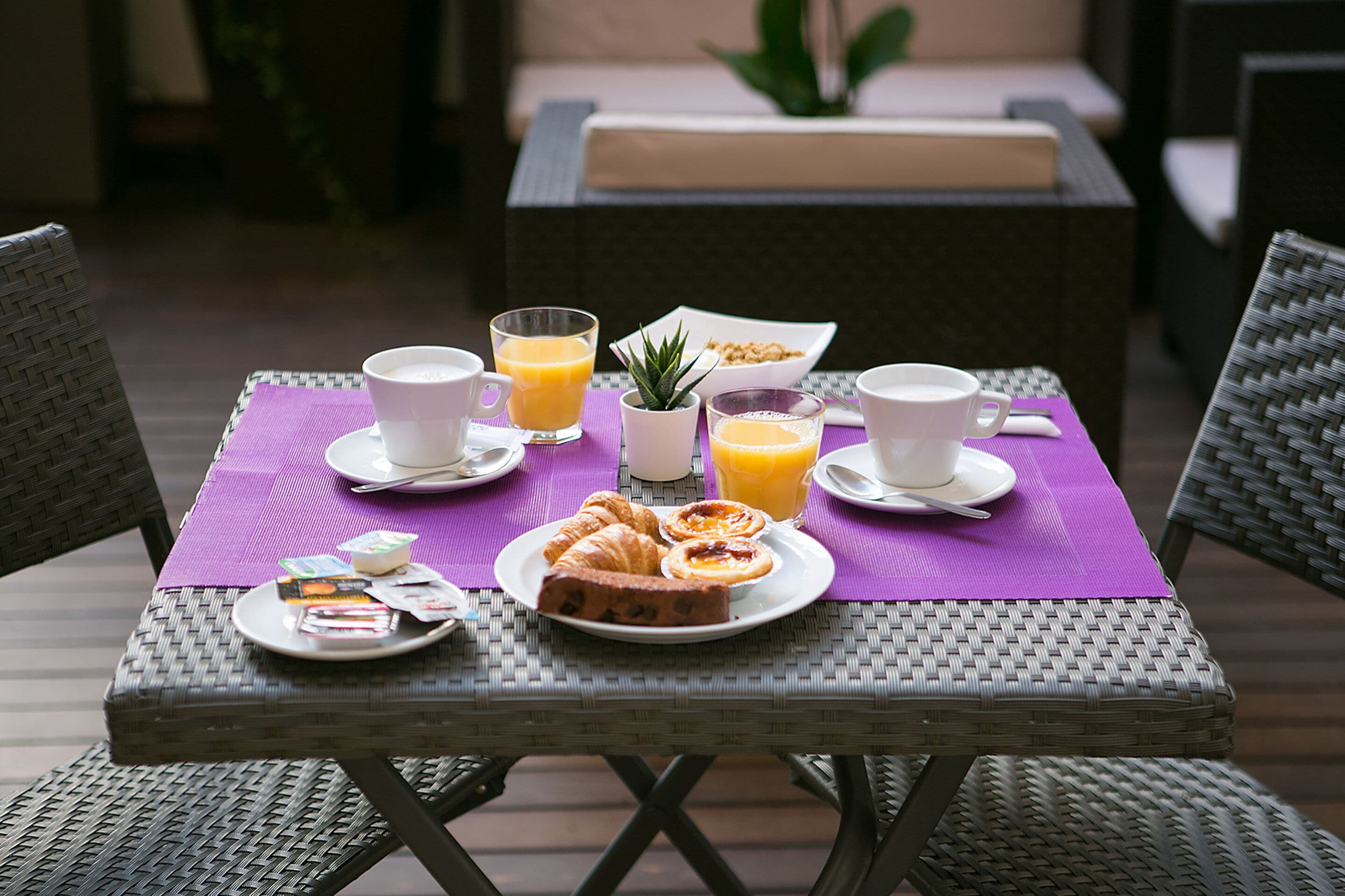 Een rieten tafeltje gedekt met ontbijtartikelen, zoals croissants, sap en koffiekopjes, met zicht op een loungehoek op de achtergrond.
