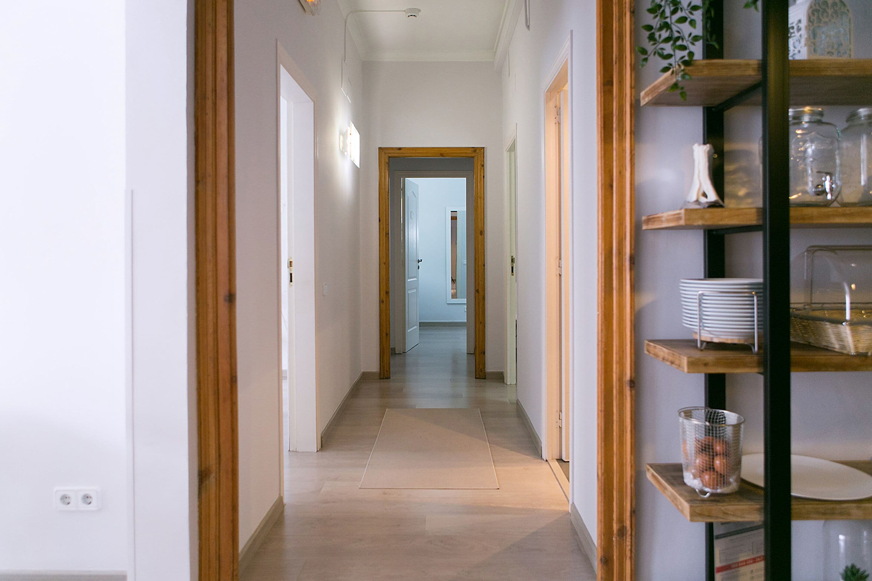 A hallway with wooden doors, a shelf with kitchenware, and a view into another room.