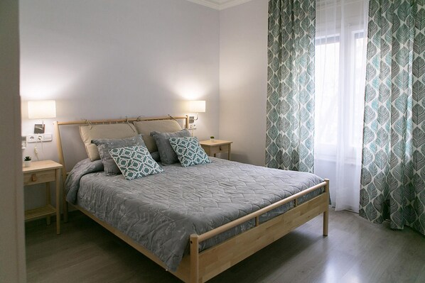 A single bed with a wooden frame, two bedside tables, a lamp on each, patterned curtains, and a small plant on the left table. - Blanc Guesthouse (Barcelona)