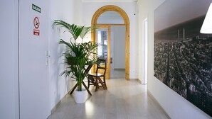 A hallway with a white door marked "SALIDA" and a "No Smoking" sign, a potted plant, a wooden chair, and a cityscape mural on the wall. - Blanc Guesthouse (Barcelona)