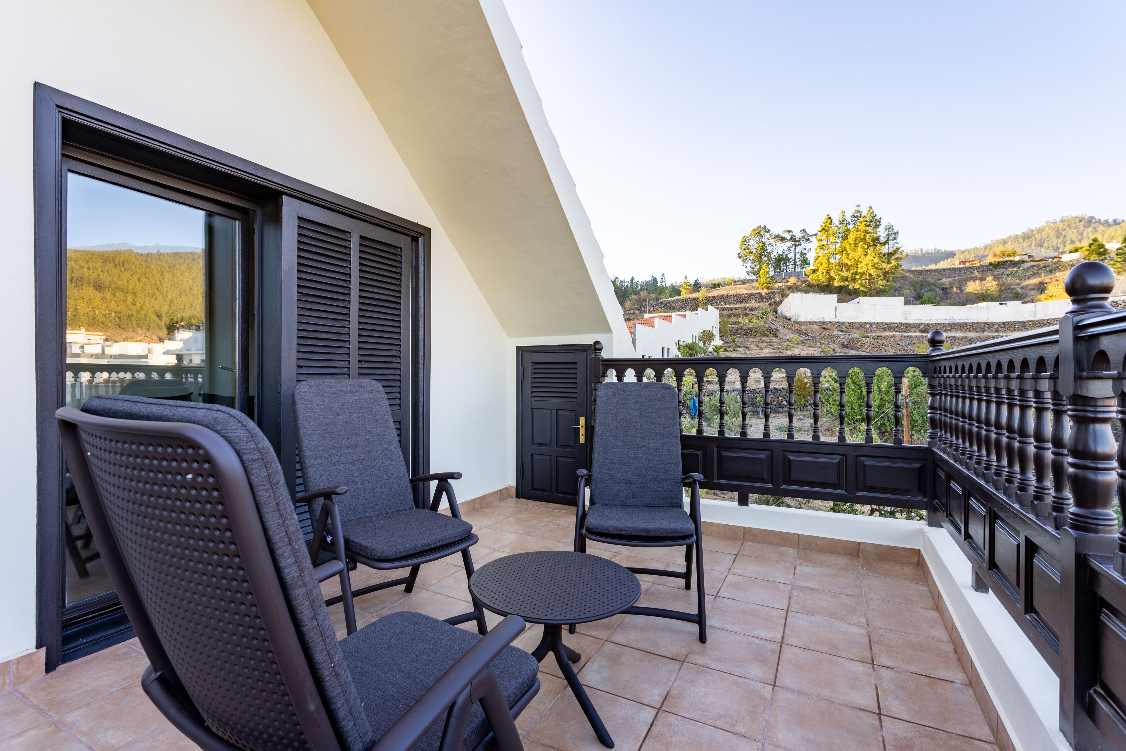 A balcony with two chairs and a small table, a black railing, and a view of a hilly landscape.