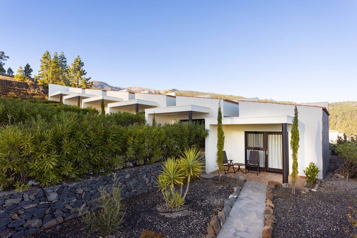 A modern building with multiple balconies, surrounded by lush greenery and a stone wall.