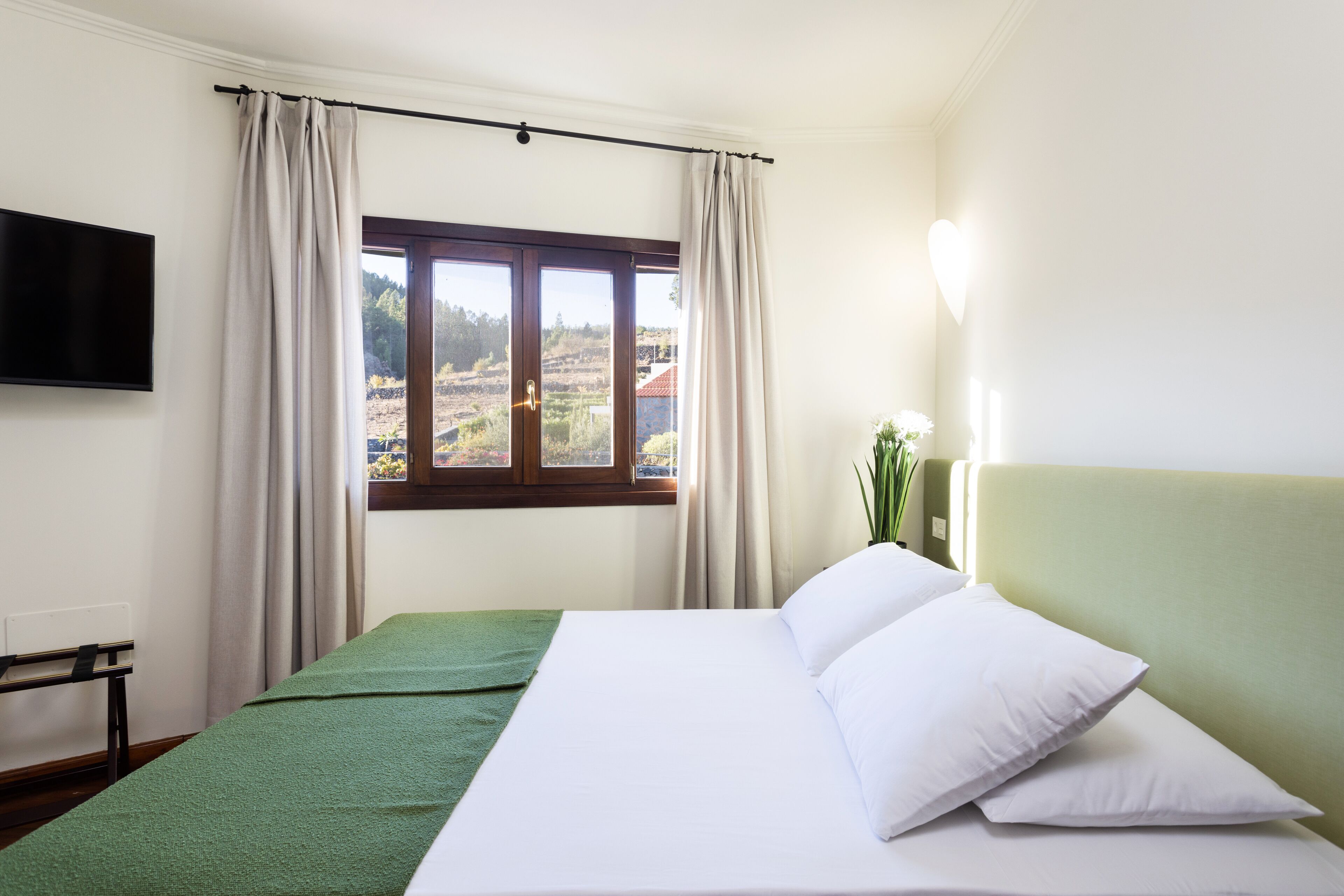 Une chambre d’hôtel avec un lit, un téléviseur fixé au mur, une fenêtre avec des rideaux et une vue sur le paysage environnant.