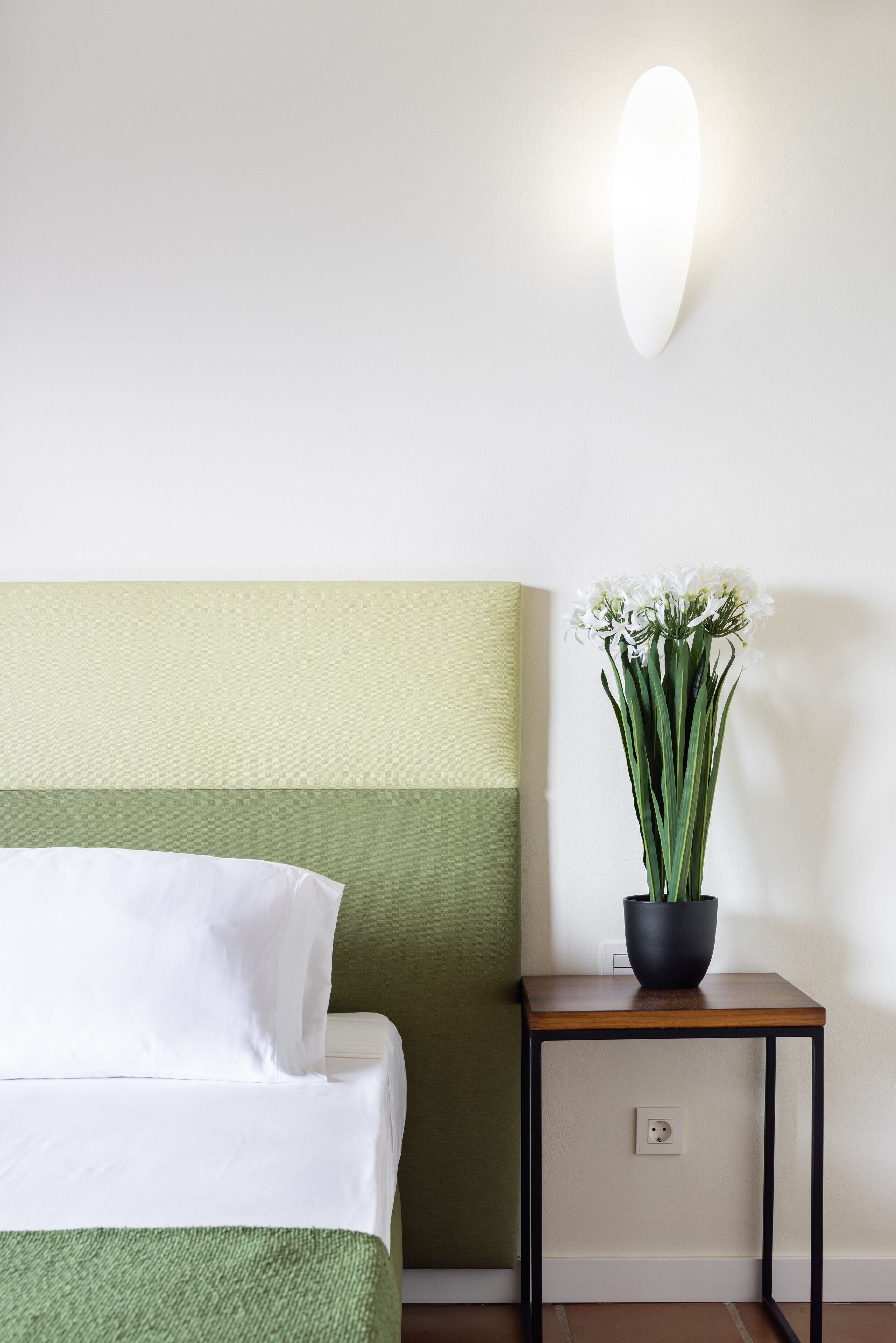 A neatly made bed with white linens, a green headboard, and a small table with a potted plant beside it.
