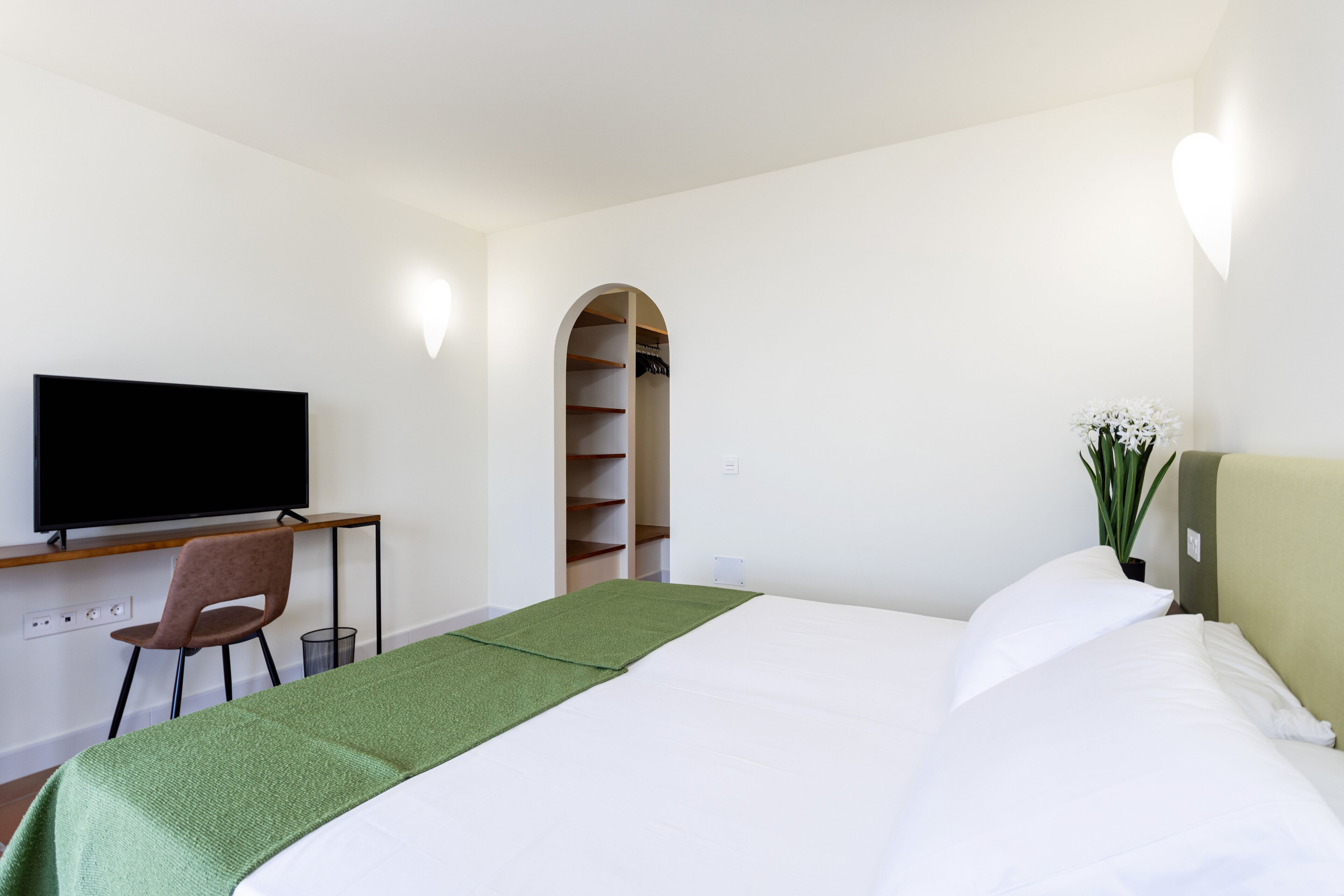 Een hotelkamer met een bed, een bureau met stoel, een televisie, een kledingkast en een plant.