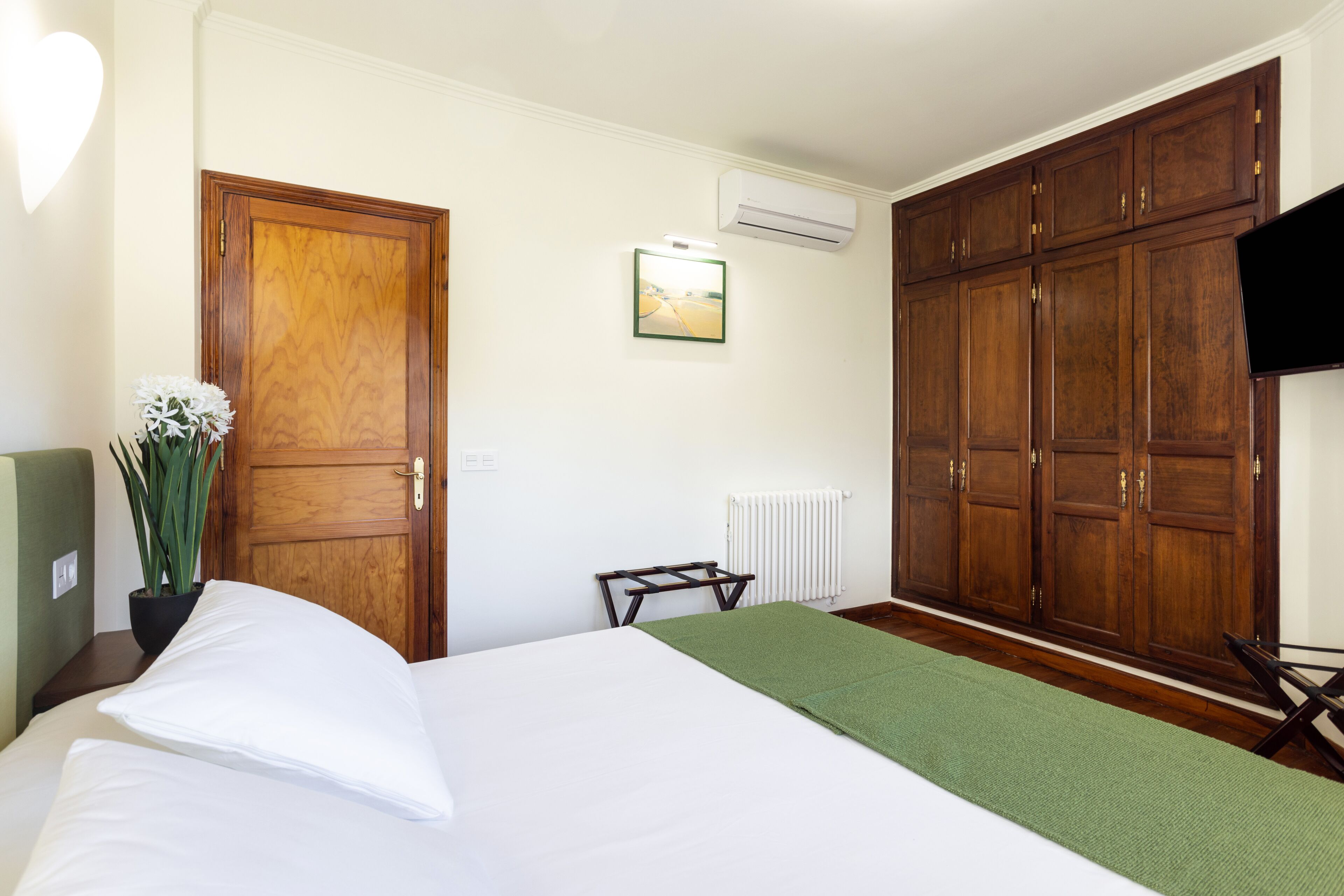 Una habitación de hotel con una cama, un plaid verde, un armario de madera, un radiador y un televisor.